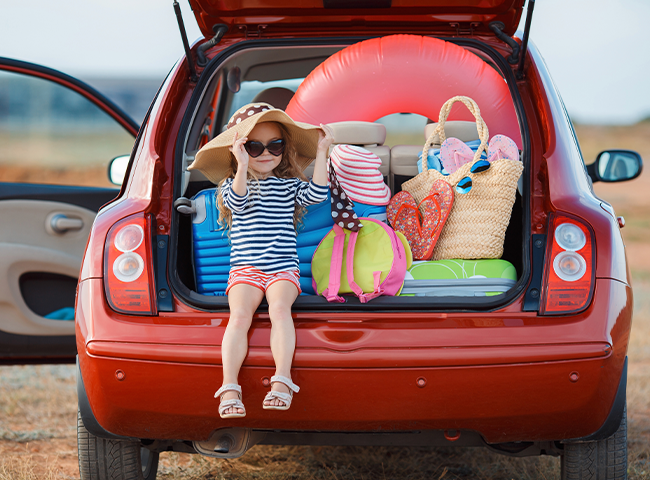 Uma menina sentada na traseira de um carro vermelho, cercada por vários itens. Ela está vestindo uma camisa listrada e um chapéu e parece estar aproveitando o tempo no carro. O carro está cheio de bagagem, incluindo mochilas, malas e bolsas. 