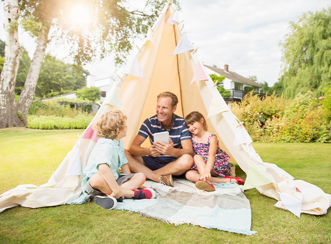 Um homem e duas crianças sentados na grama sob uma tenda. O homem está segurando um tablet, possivelmente mostrando algo às crianças. Eles estão todos sorrindo e aproveitando o tempo juntos. A barraca oferece um espaço aconchegante e confortável para a família passar bons momentos ao ar livre.