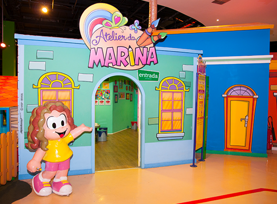 A personagem de desenho animado Marina, está parada em frente a uma casa, que parece ser um ateliê ou área de recreação infantil. Marina está posando para uma foto e parece convidar as pessoas a entrar. A casa é pintada com cores vivas, tornando-a visualmente atraente e atrativa para as crianças.