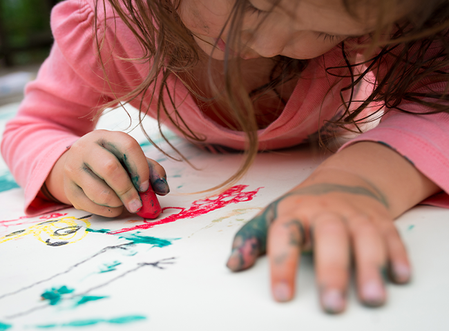 Uma menina está sentada no chão, pintando um quadro com um pincel vermelho. Ela está vestindo uma camisa rosa e parece estar gostando do processo criativo. A pintura em que ela está trabalhando está colocada sobre uma mesa à sua frente.