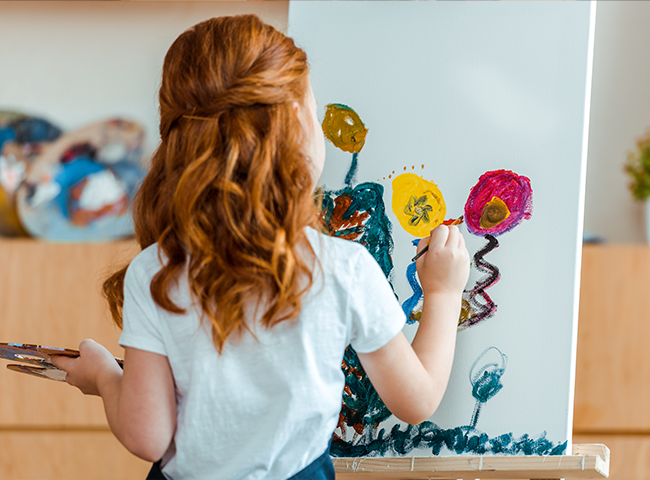 Uma menina está pintando um quadro em uma tela. Ela está vestindo uma camisa branca e está focada em seu trabalho artístico. A pintura apresenta uma árvore e balões, que ela adiciona cuidadosamente à tela. A menina está usando um pincel para aplicar a tinta, e a cena capta sua criatividade e dedicação à sua arte.