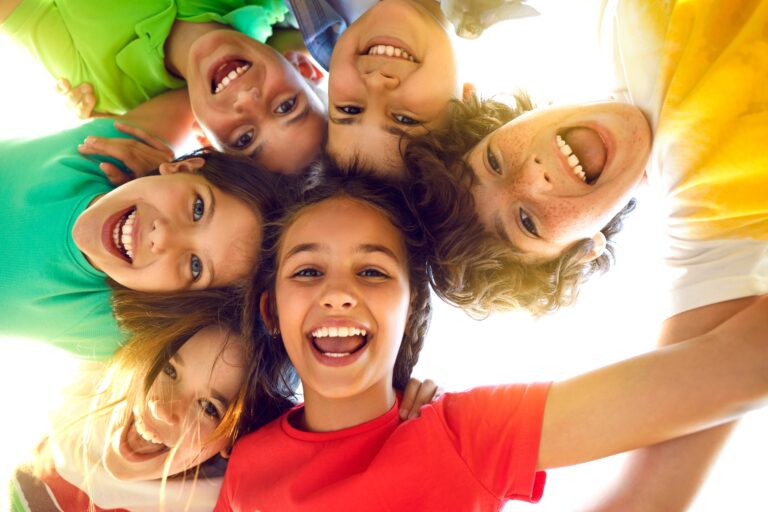 Um grupo de cinco crianças sorridentes vistas de baixo para cima, provavelmente unindo as cabeças em um círculo e olhando diretamente para baixo na direção da câmera. A luz do sol está criando um efeito de brilho que ilumina a cena, sugerindo que a foto foi tirada ao ar livre em um dia ensolarado.