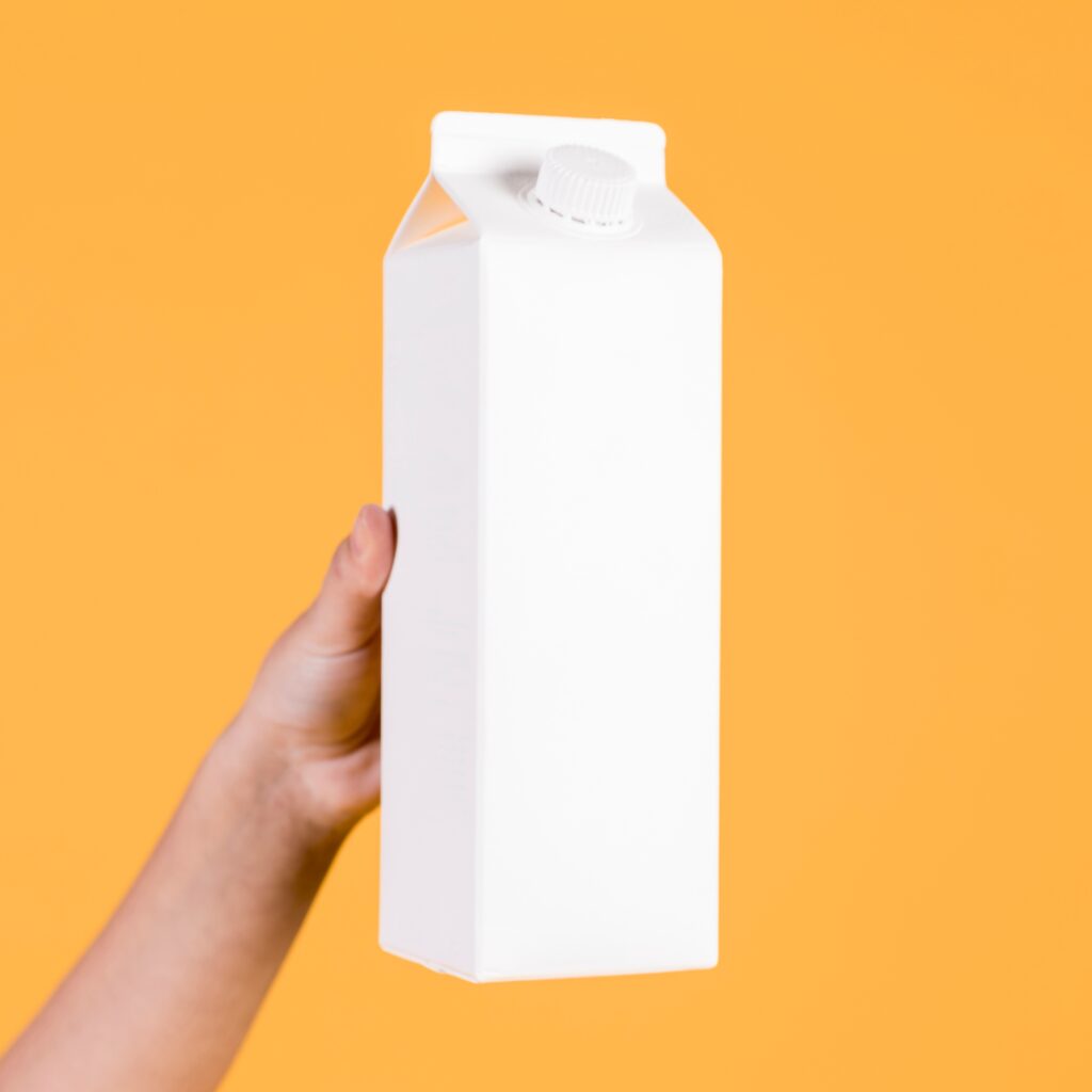 Uma mão segurando uma caixa branca com formato retangular e alongado, que tem aparência de ser uma embalagem de leite ou suco. A embalagem tem uma tampa de rosca de plástico na parte superior. O fundo da imagem é de cor laranja sólida, criando um contraste com a embalagem branca. Não há nenhuma marcação ou rótulo visível na embalagem, indicando que pode ser um mockup genérico ou uma embalagem sem marca.
