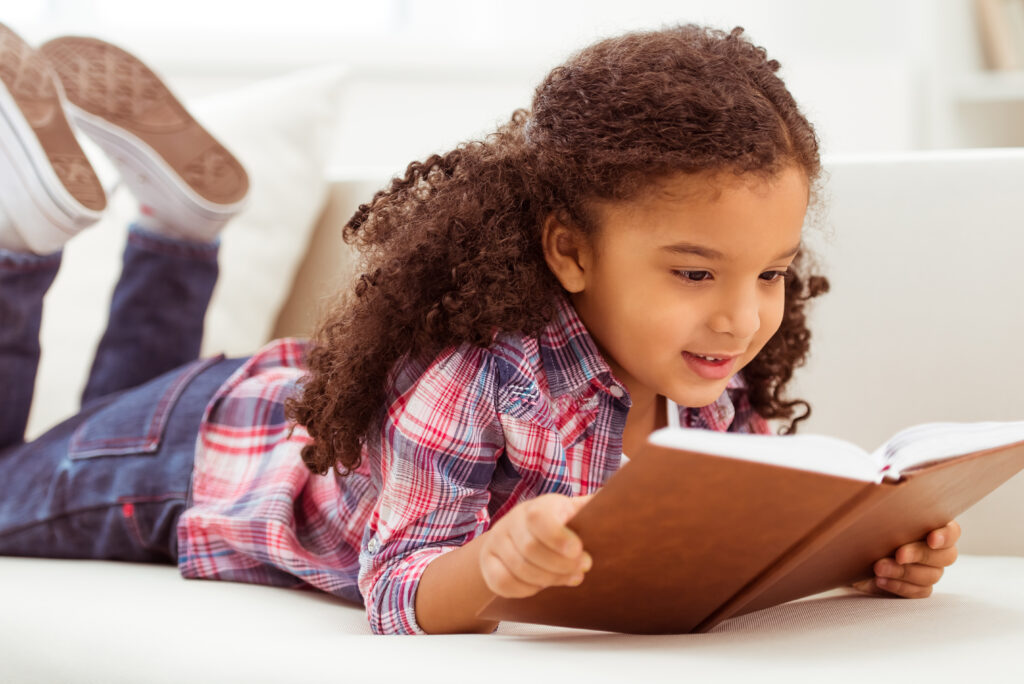 Uma menina de cabelos cacheados castanho claro está deitada em um sofá, lendo um livro. Ela está usando uma camisa xadrez e uma calça jeans, e parece estar aproveitando seu tempo. O livro está apoiado em seu colo, e ela está focada no conteúdo.

