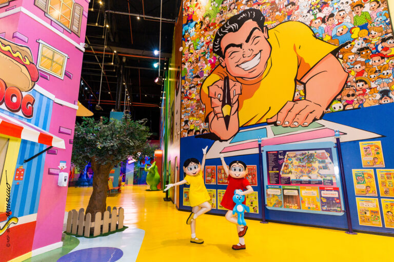 Um grande mural na parede apresenta o personagem Mauricio de Sousa em desenho animado. Abaixo, Mônica e Magali estão em frente ao mural, posando para uma foto. Ambas vestem vestidos, sendo o de Mônica vermelho e o de Magali amarelo.